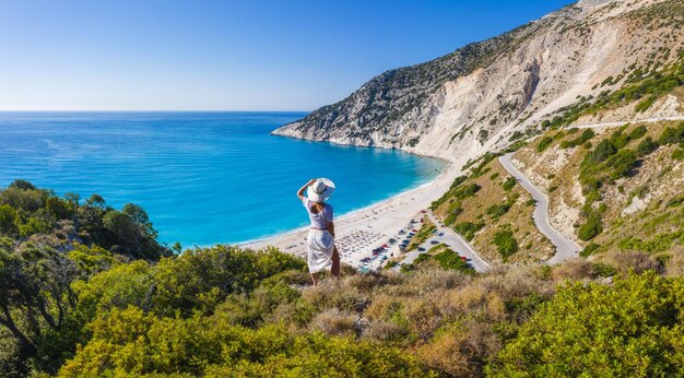 Gelukkige vrouw die bovenop een rots staat met hoed die geniet van Myrtos Beach Kefalonia eiland Griekenland