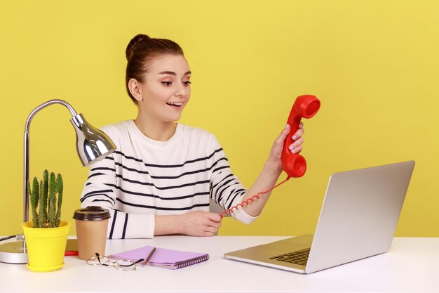 Gelukkige vrouw die een retro telefoonhandset vasthoudt en toont op het laptopscherm en vraagt om de telefoon te beantwoorden