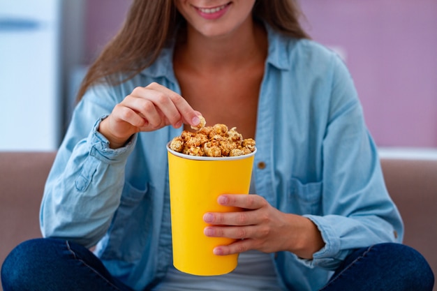 Gelukkige vrouw die en knapperige karamelpopcorn rusten eten terwijl thuis het letten op TV. Popcorn film