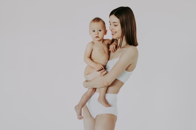 Gelukkige vrouw die haar baby op gekleurde achtergrond houdt - Jonge vrouw die ondergoed draagt dat voor haar zoontje zorgt - Zwangerschap, moederschap, mensen en verwachtingsconcept