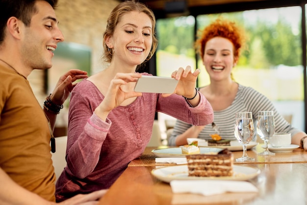 Foto gelukkige vrouw die het plakje cake van een vriend fotografeert met mobiele telefoon in een café