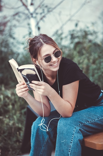 Gelukkige vrouw die naar muziek luistert met oortelefoons en een boek leest op straat