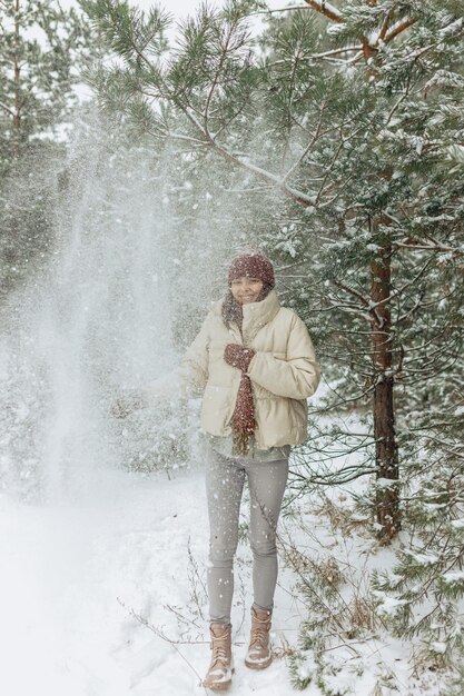 Gelukkige vrouw die plezier heeft in het winterbos terwijl ze geniet van sneeuw die uit de boom valt