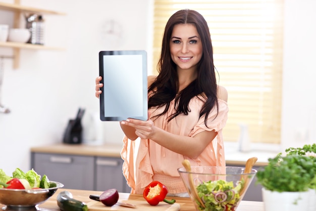 Gelukkige vrouw die salade in moderne keuken voorbereidt en tablet houdt