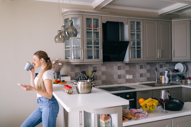 Gelukkige vrouw drinkt thuis thee in de keuken en houdt de telefoon vast