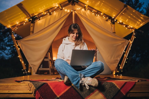 Gelukkige vrouw freelancer met behulp van een laptop op een gezellige glamping tent in een zomeravond Luxe camping tent voor buiten vakantie en vakantie Lifestyle concept