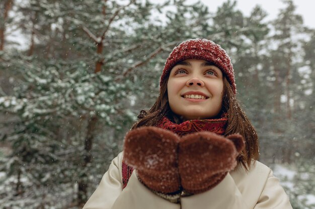 Gelukkige vrouw geniet van sneeuwval in het winterbos terwijl ze omhoog kijkt met een glimlach