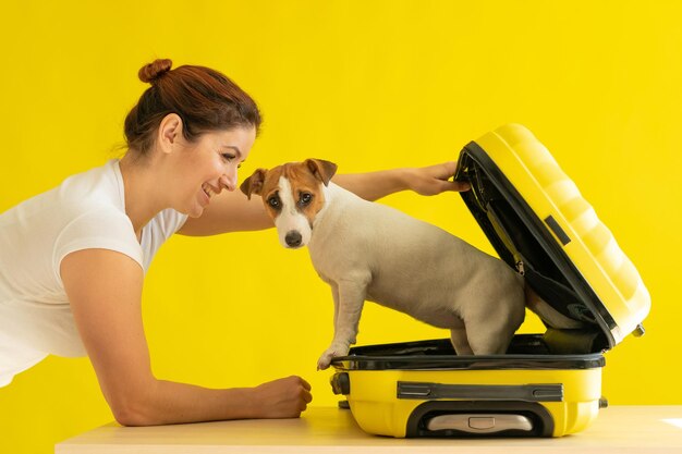 Gelukkige vrouw houdt een open koffer met een hond erin op een gele achtergrond Jack Russell Terrier puppy zit in een bagagezak en is klaar om te reizen glimlachend meisje