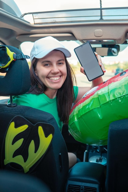 Gelukkige vrouw in auto vol met zeevakantiespullen met telefoon met witte schermkopieerruimte