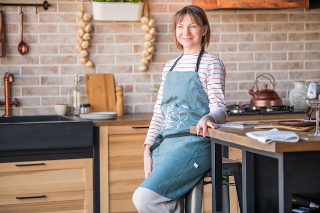 Gelukkige vrouw in een linnen schort staande in de keuken aan de tafel met een glimlach kijkt uit het raam