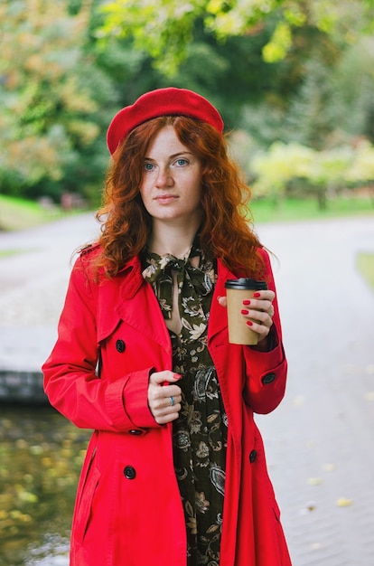 Gelukkige vrouw in een rode baretjas die in het herfstpark loopt en koffie drinkt