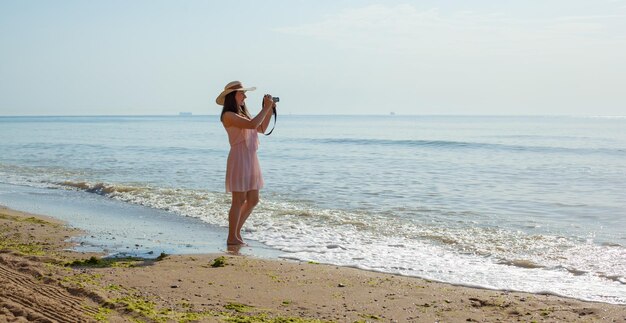 Gelukkige vrouw in een roze jurk strohoed die op de golven van de zee staat en foto's maakt met een kleine camera Ontspannende onvergetelijke vakantie op zee in de zomer Genieten van de zomer Spelen in water