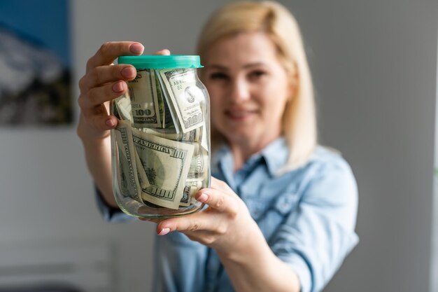 Gelukkige vrouw in grijze trui met bank met dollar contant geld, camera kijken, geïsoleerd op beige achtergrond.