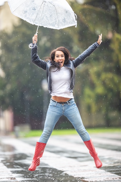 Gelukkige vrouw in rubberen laarzen en een paraplu springt van vreugde op een regenachtige dag buitenshuis.
