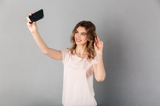 Gelukkige vrouw in t-shirt selfie maken op smartphone terwijl het aanraken van haar haren over grijs