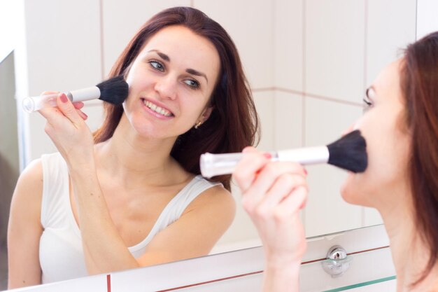 Gelukkige vrouw in wit overhemd poederend met borstel voor de spiegel in haar bordeauxrode badkamer