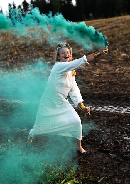Gelukkige vrouw in witte jurk met groene rookbom die op een modderig veld danst