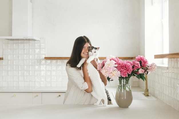Gelukkige vrouw knuffelen schattige kat op pioenrozen in vaas op achtergrond van minimale witte keuken in nieuw modern huis Huisvrouw versieren huis met bloemen en spelen met kattenhuishouding