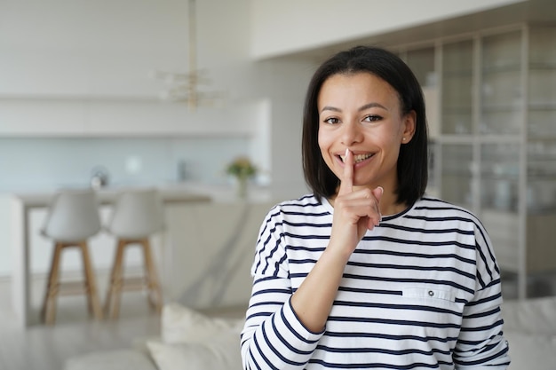 Gelukkige vrouw maakt stiltegebaar en vraagt om stil te zijn om thuis naar de camera te kijken