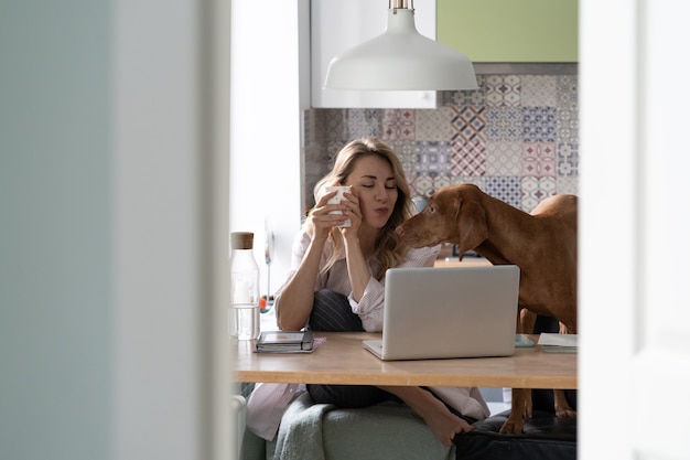 Gelukkige vrouw maakt videogesprek met echtgenoot op zakenreis in de keuken samen met hond die koffie drinkt