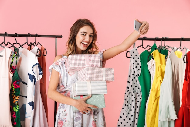 gelukkige vrouw met aankoop staan in de winkel in de buurt van kledingrek en nemen selfie op mobiele telefoon geïsoleerd op roze