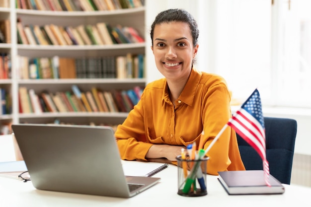 Gelukkige vrouw met de vlag van de V.S. met behulp van laptop die Amerikaans Engels online studeert, in de bibliotheek zit en naar de camera glimlacht