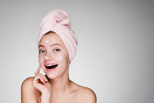 Gelukkige vrouw met een handdoek op haar hoofd bracht een crème op haar gezicht aan op een grijze achtergrond