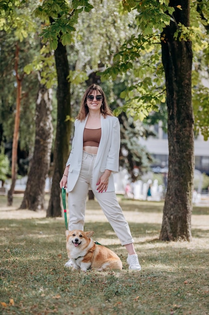 Gelukkige vrouw met een wit pak en een zonnebril die in het stadspark loopt met de Welsh Corgi Pembroke-hond