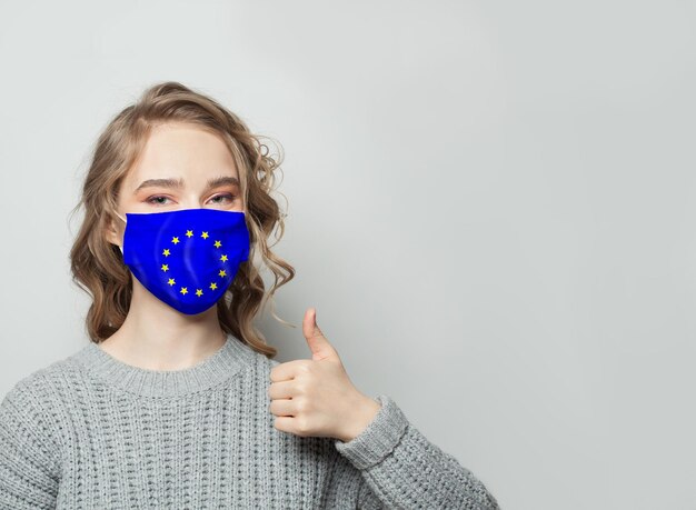 Foto gelukkige vrouw met gezichtsmasker die de duim omhoog houdt met de vlag van de eu op de achtergrond concept voor griepepidemie en virusbescherming