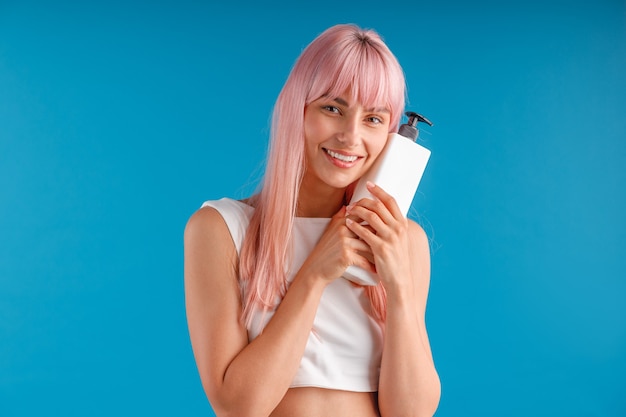 Gelukkige vrouw met glad roze haar die pompfles vasthoudt en knuffelt met haarmasker of shampoo poseren