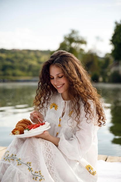 Gelukkige vrouw met krullend haar geniet van een picknick aan het meer in de buitenlucht, eet een croissant Heerlijke en gezonde picknick in de natuur