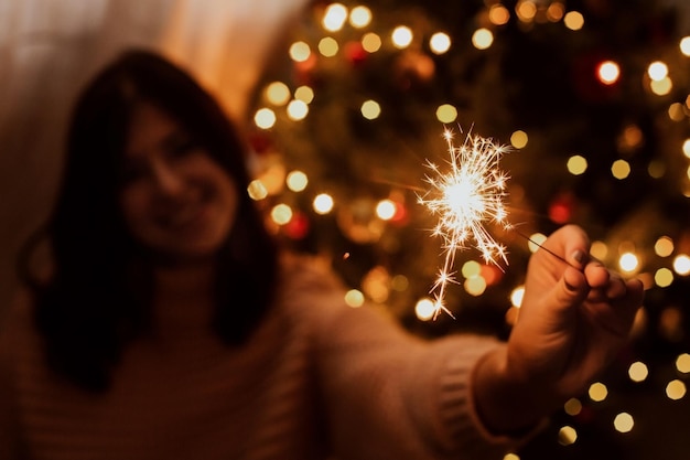 Gelukkige vrouw met vuurwerk bij kerstboom met gouden lichten Stijlvol meisje met brandend sterretje vieren in feestelijke donkere kamer Gelukkig Nieuwjaar