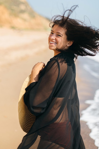 Gelukkige vrouw met winderig haar en hoed wandelen en glimlachen op zandstrand op zee Zomervakantie