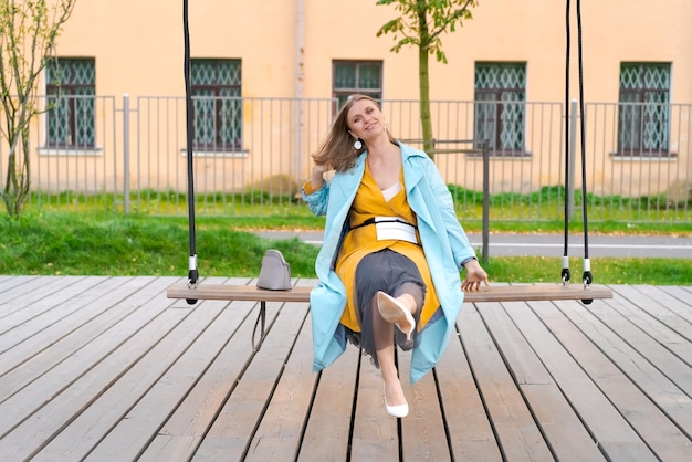 Gelukkige vrouw schommelend op schommel in het stadspark met gele jurk en blauwe jas het concept van vrijheid