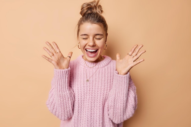 Foto gelukkige vrouw schreeuwt van vreugde sluit ogen en houdt handpalmen omhoog blij om uitstekend nieuws te horen heeft haar verzameld in knot draagt gebreide trui geïsoleerd over beige achtergrond menselijke emoties concept