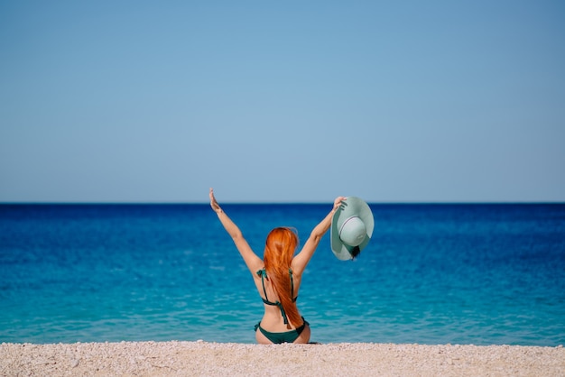 Gelukkige vrouw steekt enthousiast haar handen omhoog zittend op het strand, zonnige dag