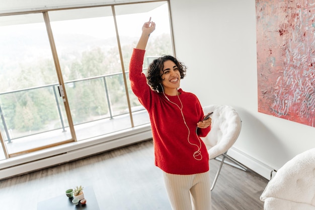 Foto gelukkige vrouw uit het midden-oosten met een mobiele telefoon die danst en naar haar favoriete liedje luistert in de woonkamer gelukkige vrouw die thuis tijd doorbrengt