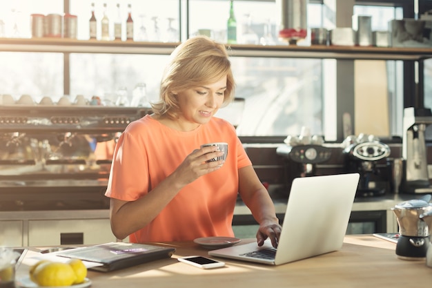 Gelukkige vrouw van middelbare leeftijd barista of manager die op laptop werkt, online een bestelling plaatst en koffie drinkt in moderne cafetaria. Werk, levensstijl, café, restaurantconcept