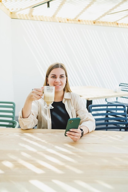 Gelukkige vrouw zit aan een buitencafé tafel en praat aan de telefoon met een kop koffie glimlachende vrouw geniet van telecommuting in een café of online studeren