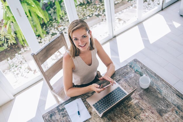 Foto gelukkige vrouw zittend aan tafel met laptop en tikken op mobiel