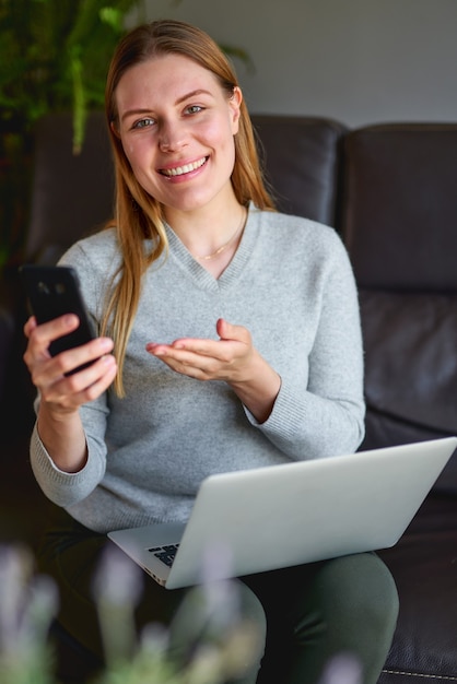 Gelukkige vrouw zittend op de bank met laptop en praten over de telefoon thuis.