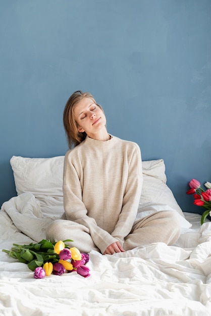 Gelukkige vrouw zittend op het bed, het dragen van pyjama's met tulp bloemen boeket, blauwe muur achtergrond