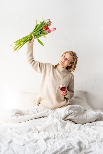 Gelukkige vrouw zittend op het bed in pyjama, met plezier genietend van bloemen en een glas rode wijn