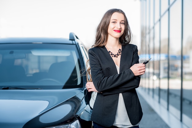 Gelukkige vrouwelijke koper met sleutels in de buurt van de auto voor het moderne avtosalon-gebouw