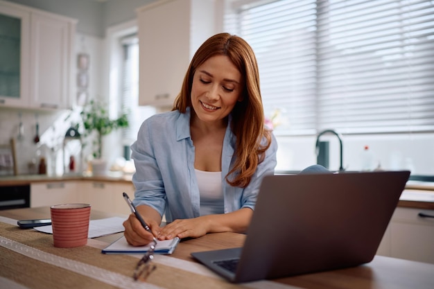 Gelukkige vrouwelijke ondernemer die thuis werkt met een laptop en in een notitieboek schrijft