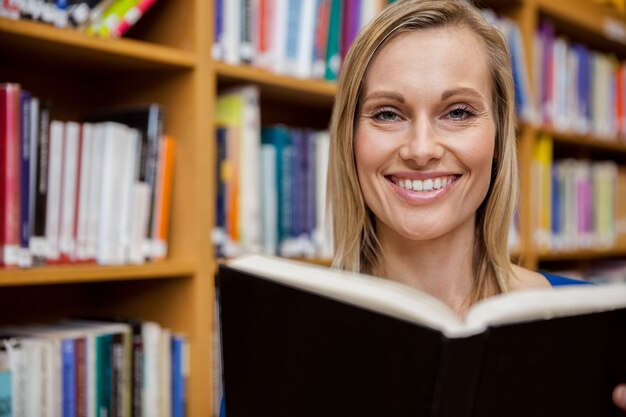 Gelukkige vrouwelijke student die een boek in de bibliotheek leest bij de universiteit