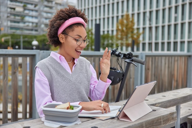 Gelukkige vrouwelijke student studeert in de verte zwaait hallo op tablet begroet iemand maakt aantekeningen in notitieboekje poses buiten tegen stedelijke achtergrond heeft videoconferentie. Studeren en technologie concept