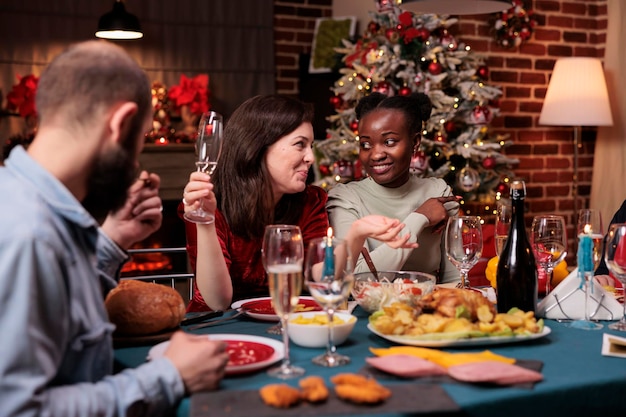 Gelukkige vrouwen die kletsen op het kerstfeest thuis, mousserende wijn drinken, wintervakantie vieren. Vrolijke vrienden praten aan feestelijke eettafel, traditioneel kerstvoedsel eten