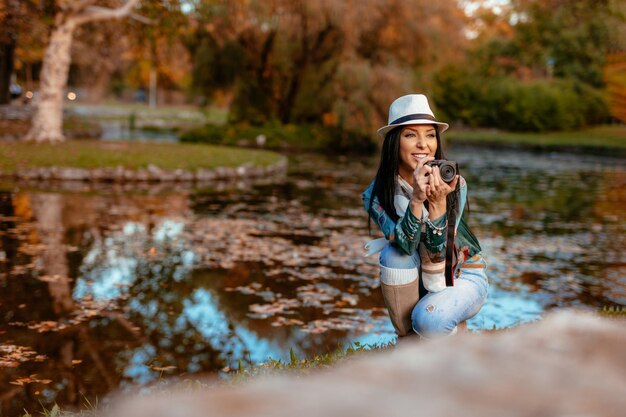 Gelukkige vrouwenfotograaf die professionele digitale camera houdt en foto neemt dichtbij het meer in het herfstpark.