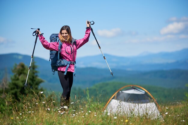 Gelukkige vrouwentoerist met rugzak en trekkingstokken dichtbij tent